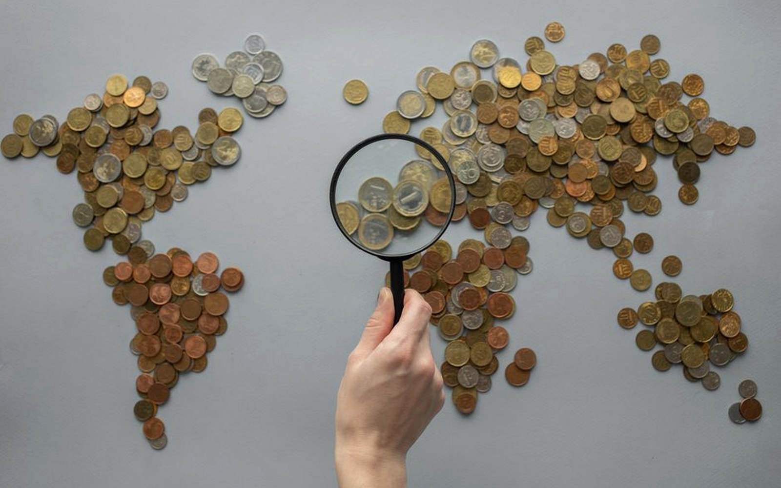 A world map assembled from coins with a magnifying glass held above it.